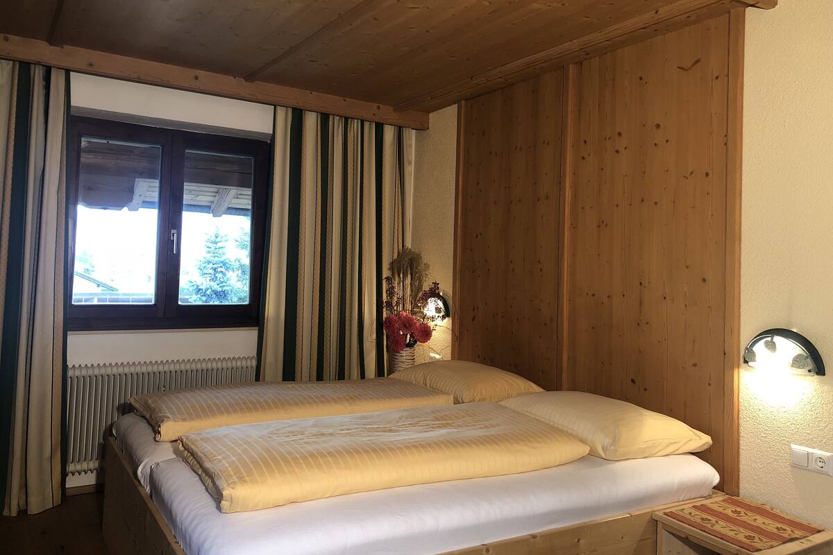 A wooden room with a bed, a window with curtains, a radiator, and a wall-mounted light fixture.