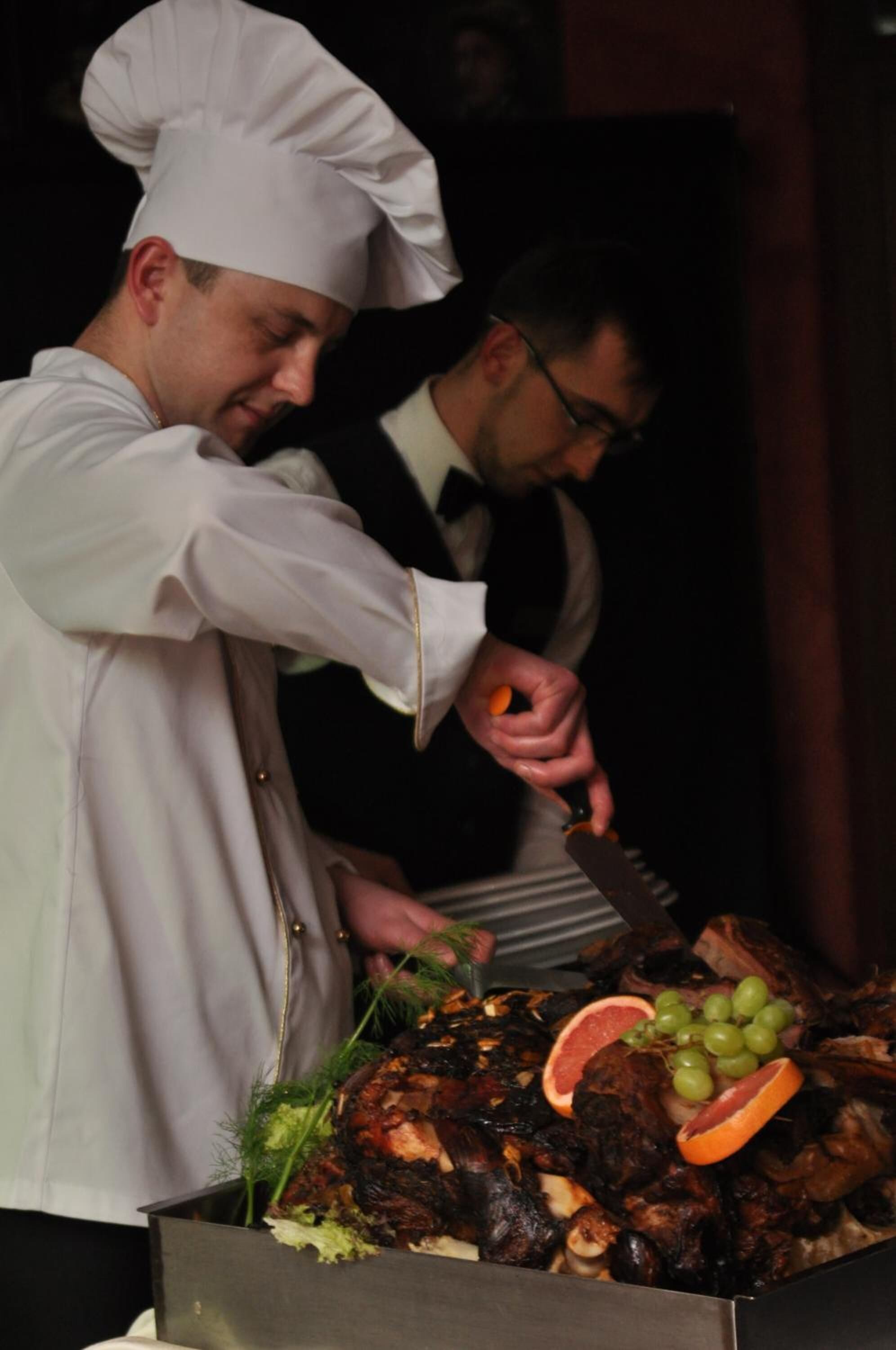 A chef slicing meat with a knife, surrounded by a variety of garnishes including grapes and citrus slices.