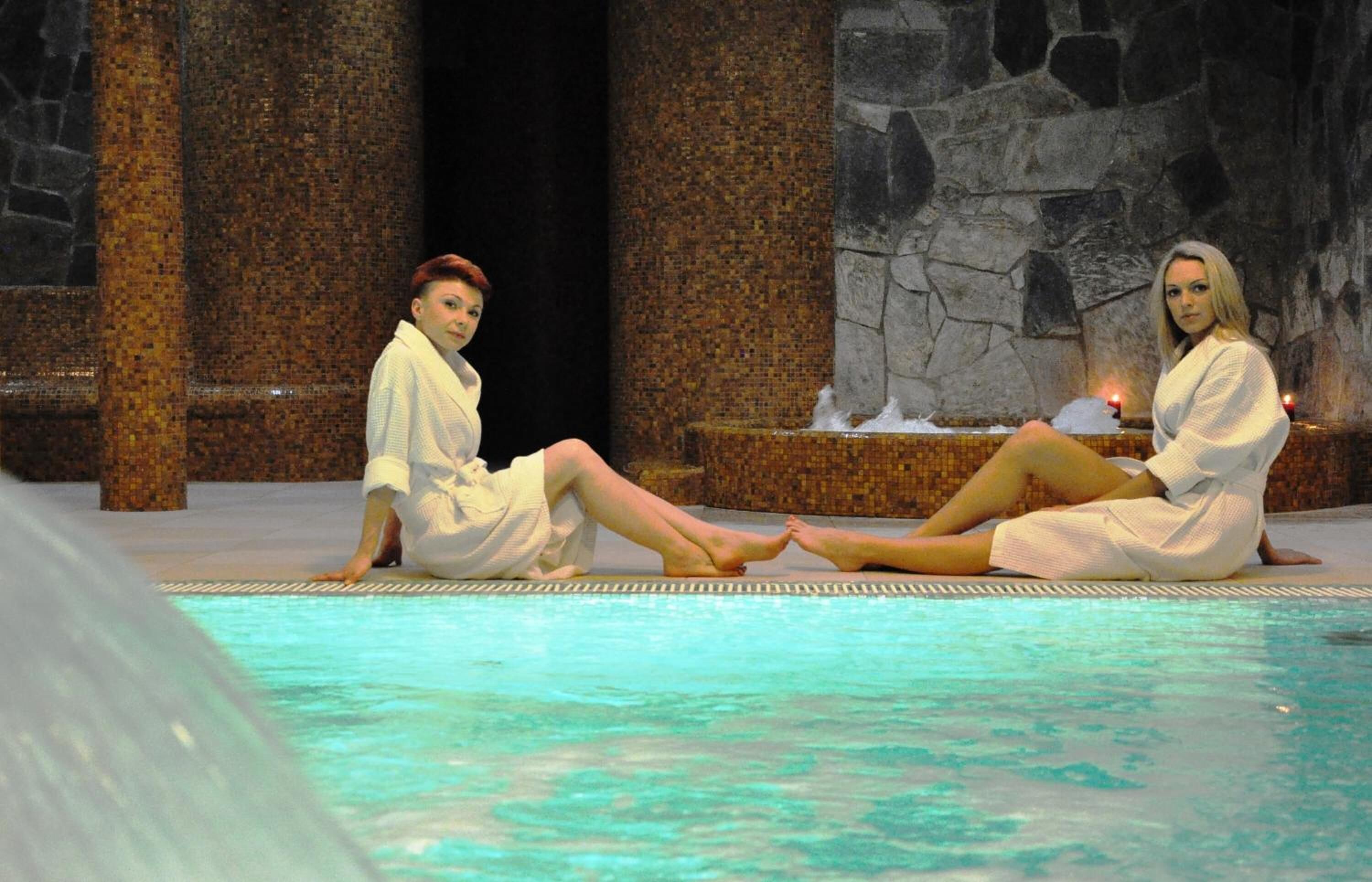 Two individuals sitting by a pool with stone columns and a tiled wall in the background.