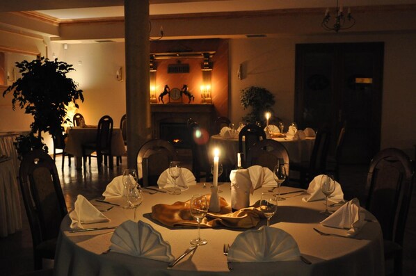 A dimly lit dining room with round tables set for a meal, candles, and a fireplace in the background. - Hotel Karino Spa (Solina)