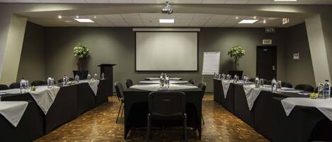 A conference room with a large screen, multiple tables with chairs, water bottles, and a podium.