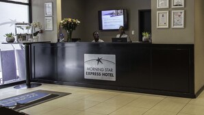 Mostrador de recepción del Hotel Morning Star Express, con premios enmarcados en la pared, una pantalla de televisión y un jarrón de flores.