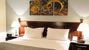 Una habitación de hotel con una cama grande, dos mesitas de noche con lámparas, y tres cuadros abstractos en la pared.