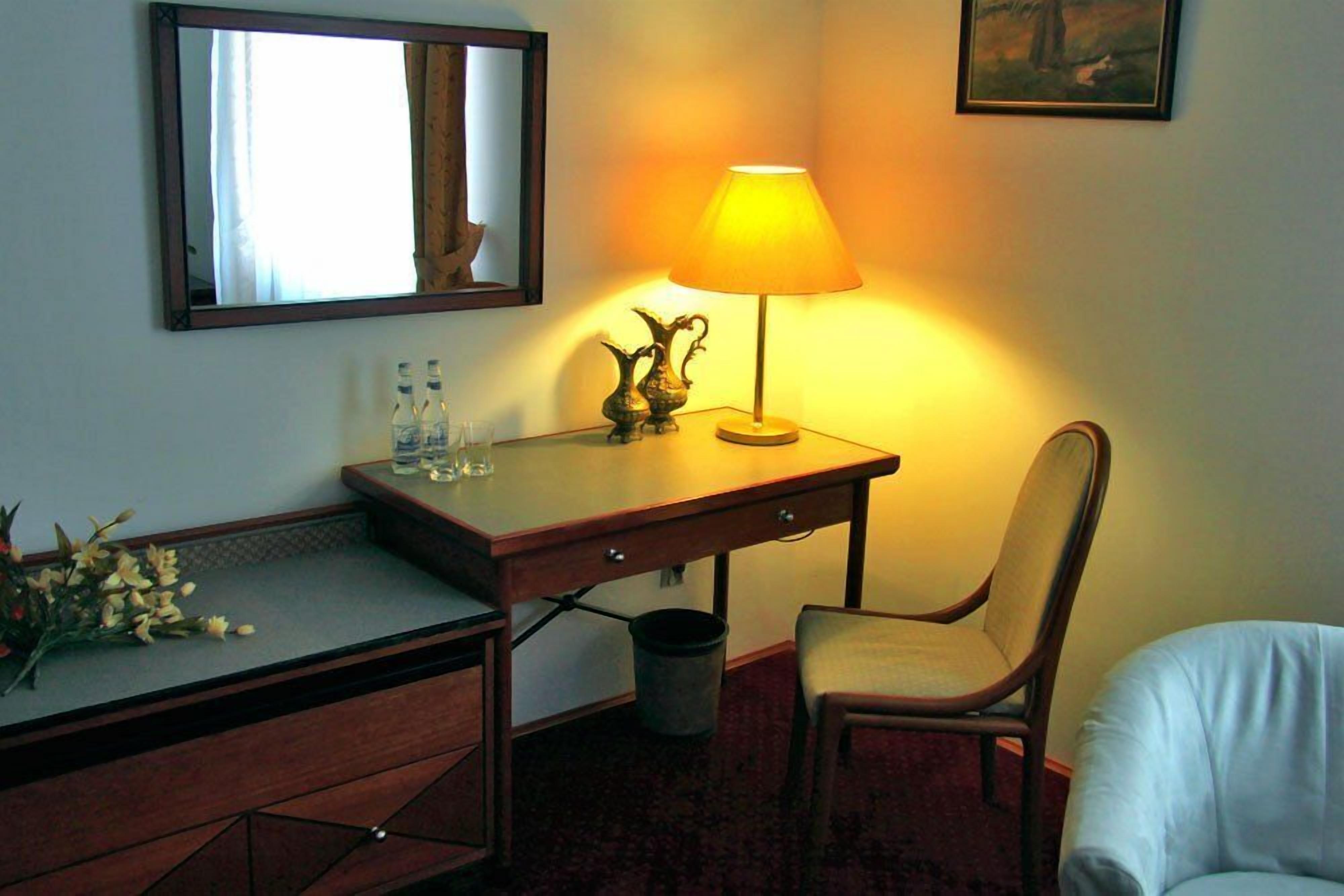 Una habitación de hotel con escritorio, silla, lámpara, espejo y un pequeño cubo de basura.