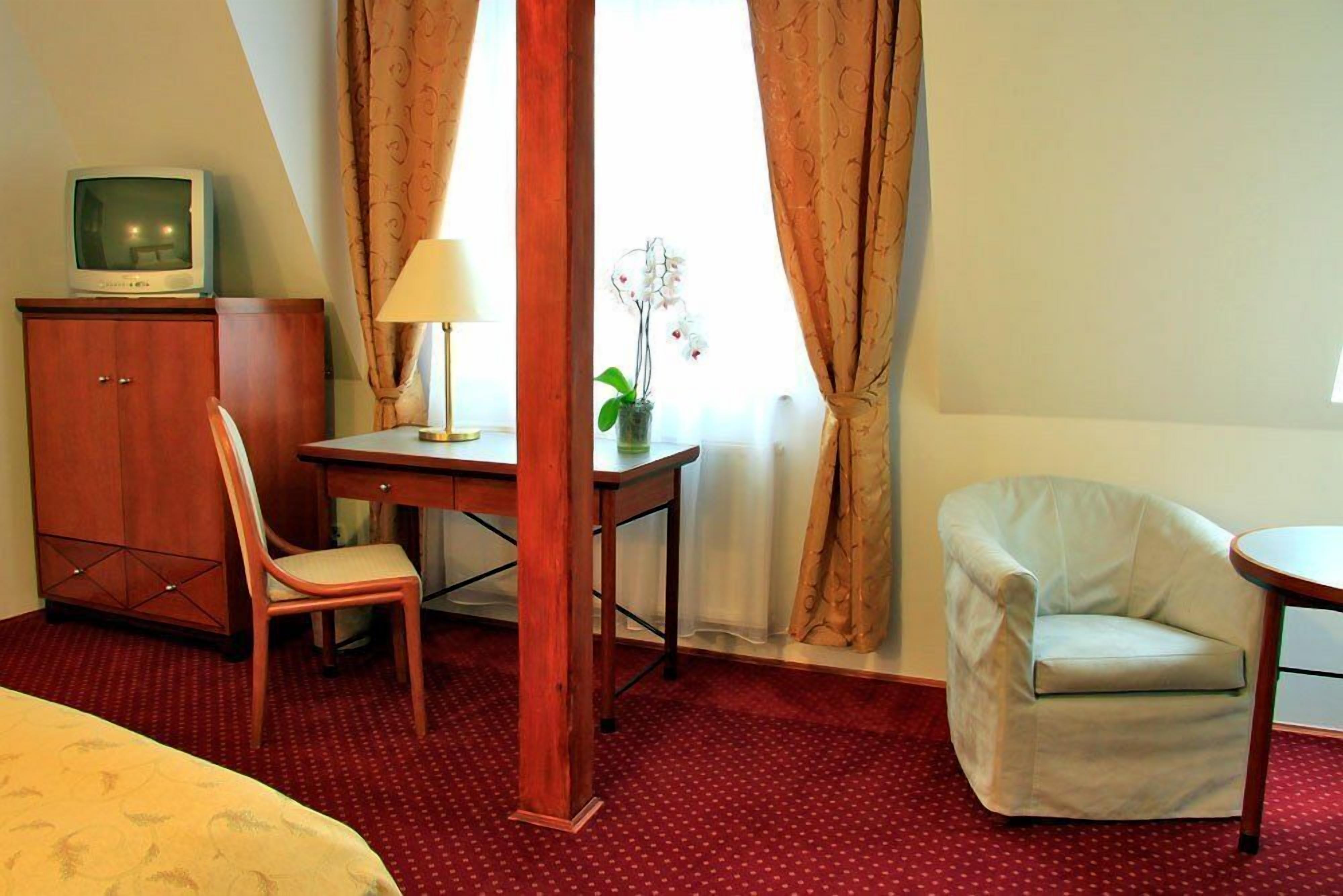 Ein Hotelzimmer mit Bett, einem Holztisch mit Lampe, einem Sessel, einem Fernseher und einem Fenster mit Vorhängen.