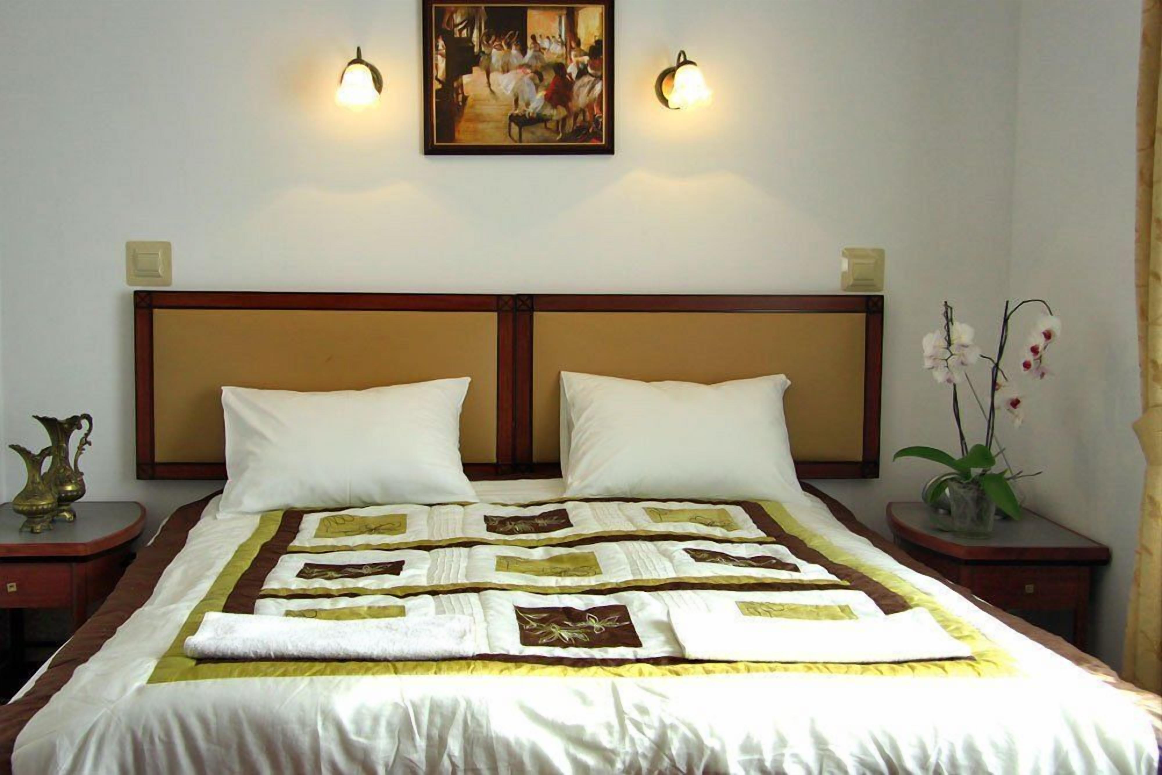 A neatly made bed with a quilt, two bedside tables with lamps, a painting on the wall, and a vase with flowers.