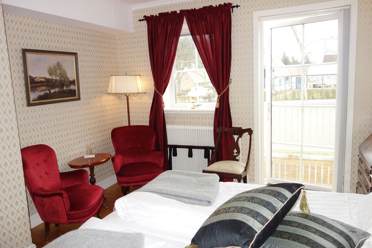 A hotel room with a bed, two red armchairs, a small table, a lamp, and a window with curtains.