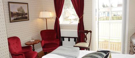 Une chambre d’hôtel avec un lit, deux fauteuils rouges, une petite table, une lampe et une fenêtre avec des rideaux.