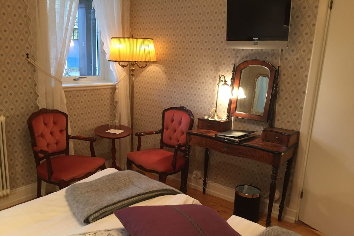 A hotel room with a bed, two red upholstered chairs, a wooden desk with a mirror, a lamp, and a television mounted on the wall.
