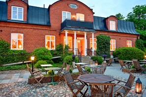 A brick building with a balcony, outdoor seating area with tables and chairs, and a well-maintained garden.