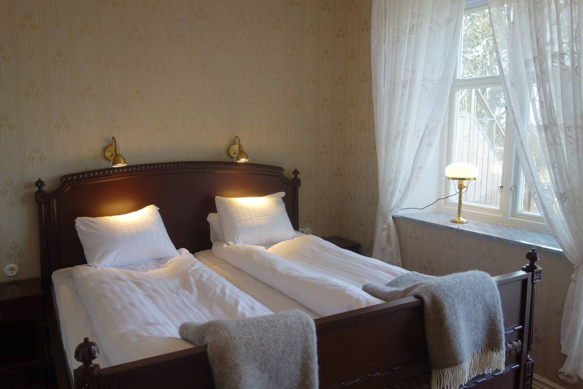 A double bed with white linens, two bedside lamps, and a window with sheer curtains.