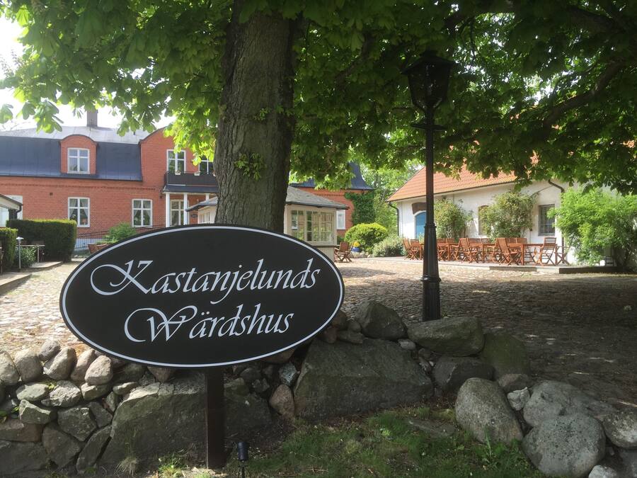 Señal de Kastanjelunds Vårdsby, un edificio de madera tradicional con muros de piedra y un camino adoquinado.