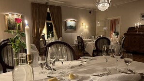 En elegant spisesal med dækkede borde, lysekroner, indrammede kunstværker og udsigt til det fri.