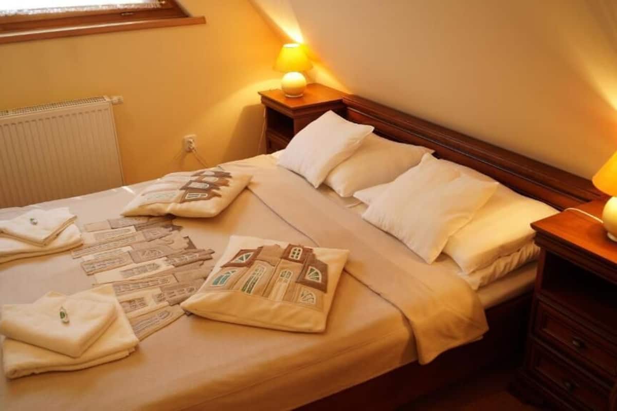 Ein ordentlich bezogenes Bett mit Kissen und Decke, ein Nachttisch mit Lampe und ein Fenster mit Jalousien.