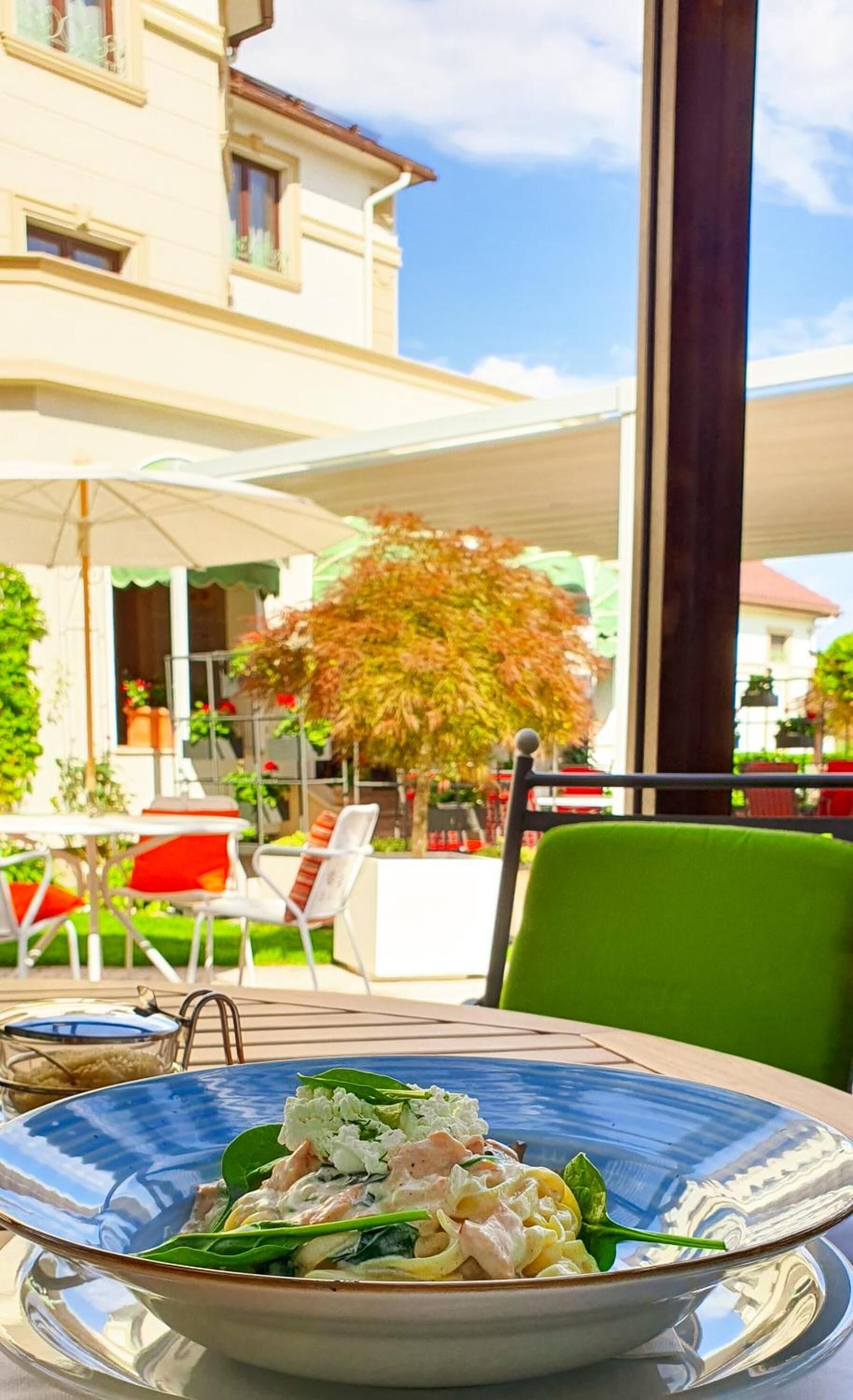Un área de comedor al aire libre con una mesa dispuesta para una comida, rodeada de sillas y un edificio al fondo.