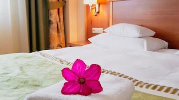 A neatly made bed with white linens and a pink flower on top, a wall-mounted lamp, and a painting above the bed.