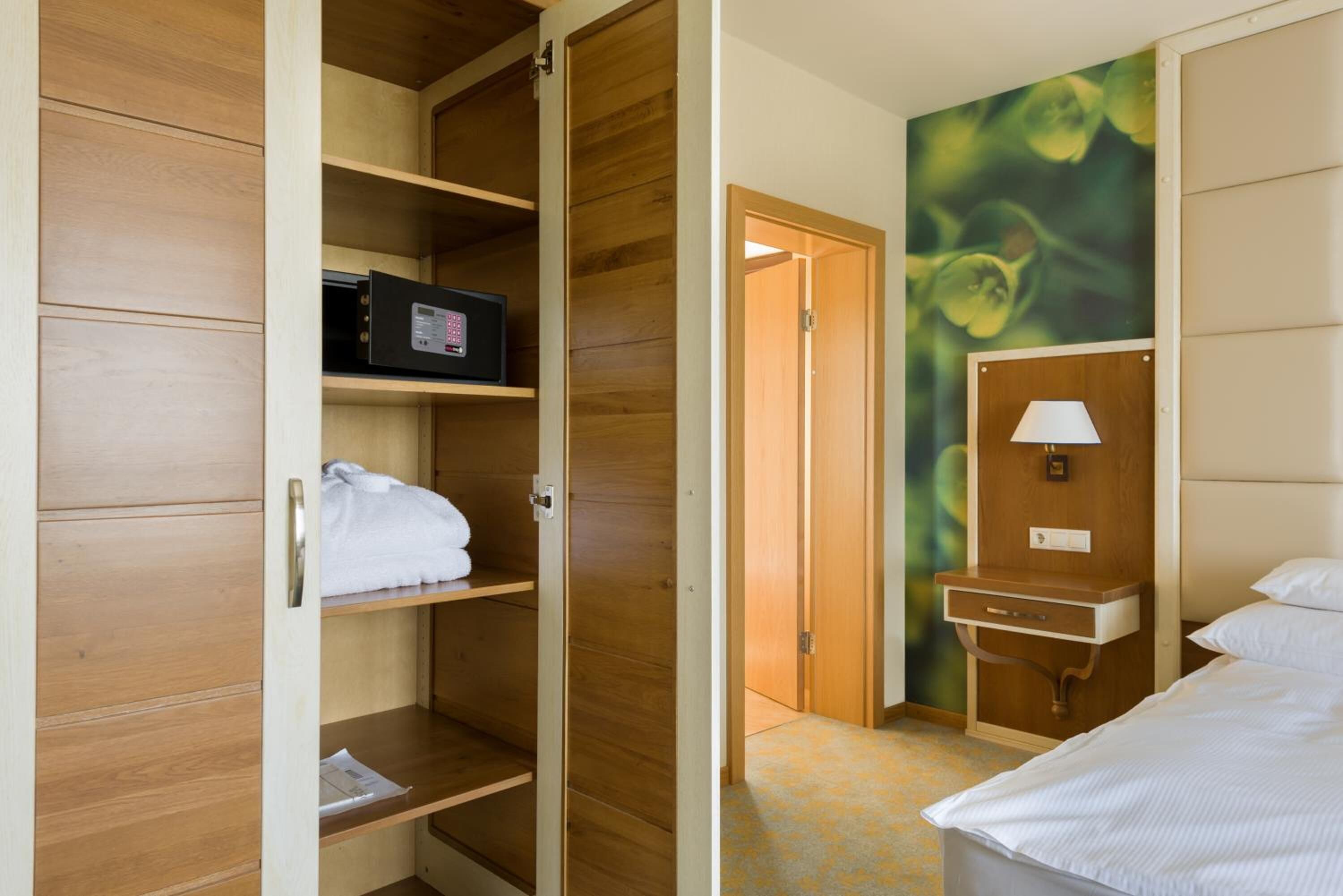 A hotel room with a wooden wardrobe, a bed, a bedside table with a lamp, and a wall with a green abstract design.