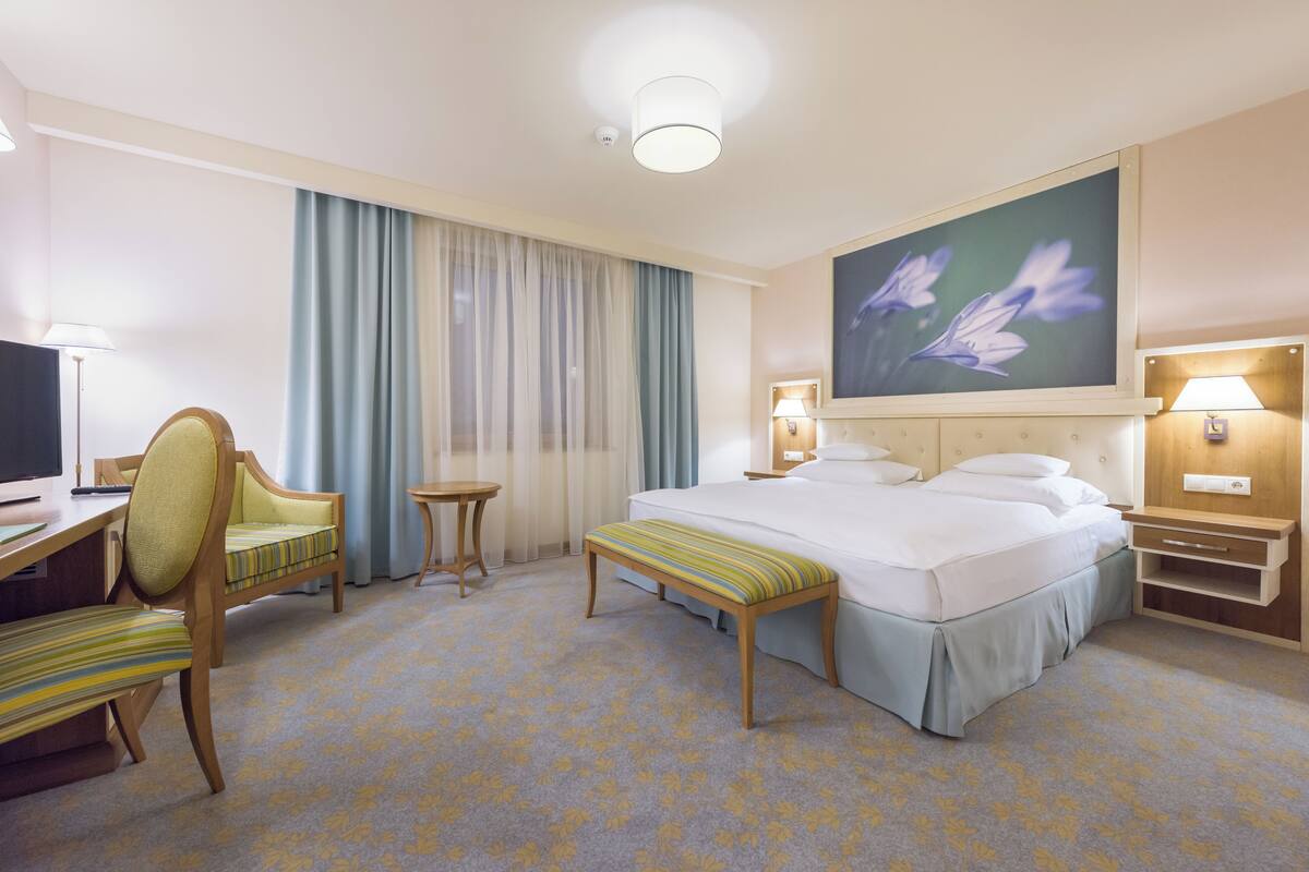 A hotel room with a bed, a desk, a chair, a small table, and a painting on the wall.