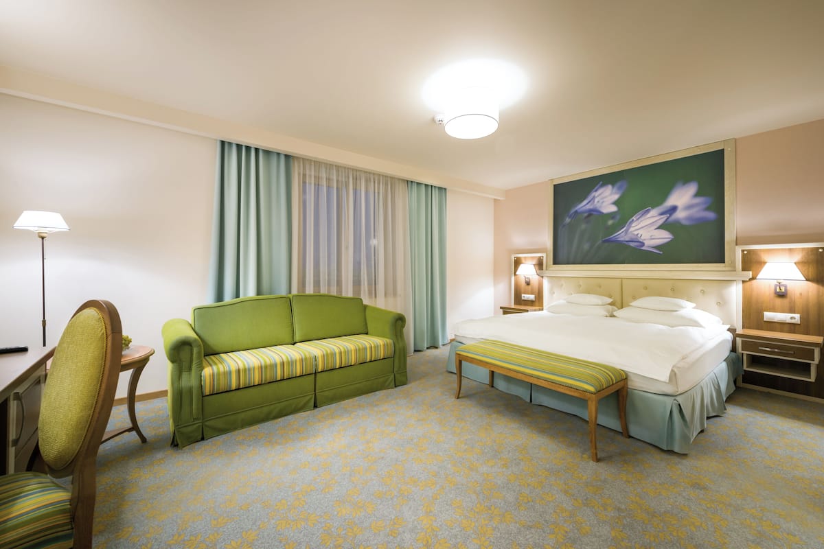 A hotel room with a bed, a green sofa, a wooden bench, a desk with a lamp, and a painting on the wall.