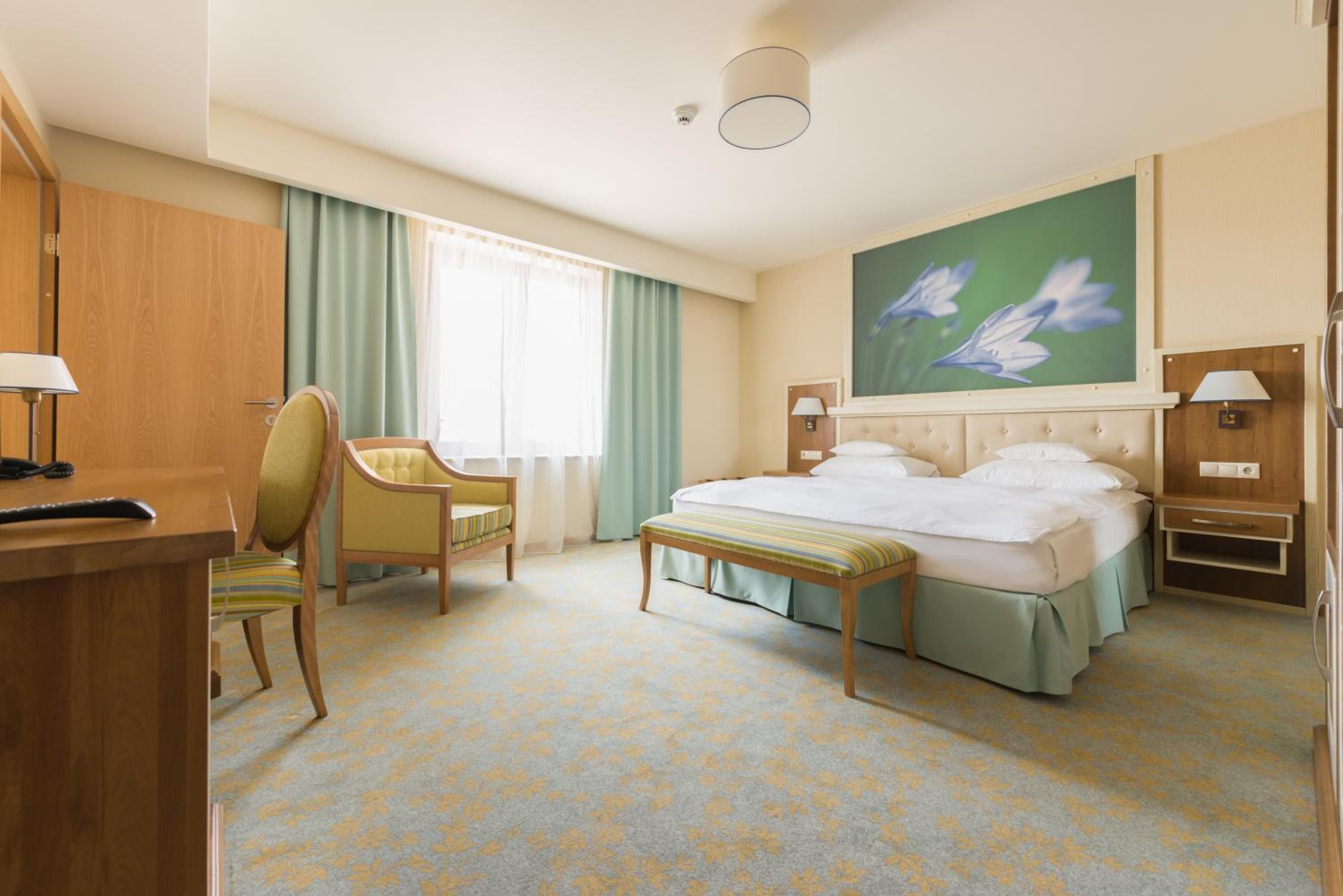 A hotel room with a bed, a desk, a chair, a bench, and a painting of flowers on the wall.