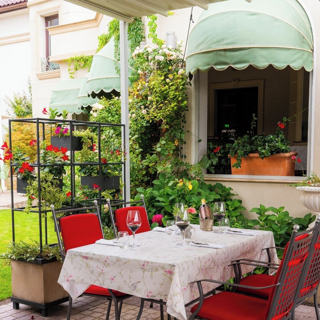 An outdoor dining area with a table set for two, surrounded by red chairs, a metal plant stand with flowers, and a green awning over a window.