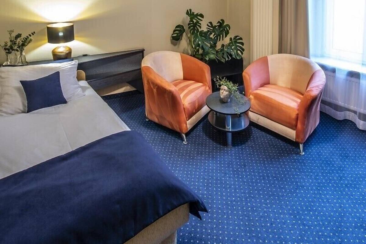 A hotel room with a bed, two armchairs, a small table, a lamp, a potted plant, and a window with curtains.