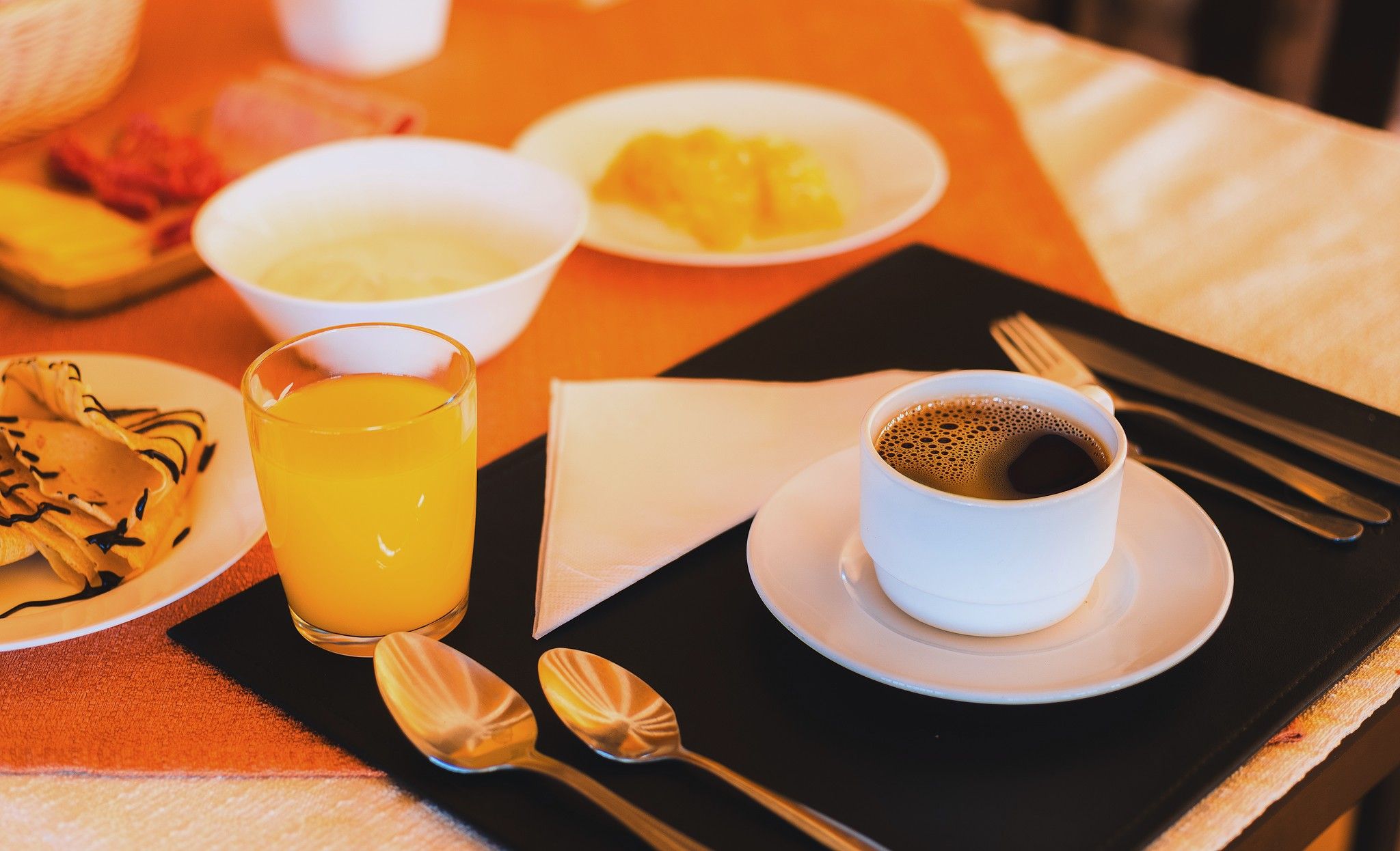 Une table mise pour le petit-déjeuner avec un verre de jus d’orange, une tasse de café et des assiettes remplies de nourriture, notamment des crêpes et des fruits.