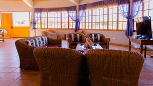 Un salón amplio con mobiliario de mimbre, ventanas grandes con cortinas moradas y un televisor sobre una mesa de madera.