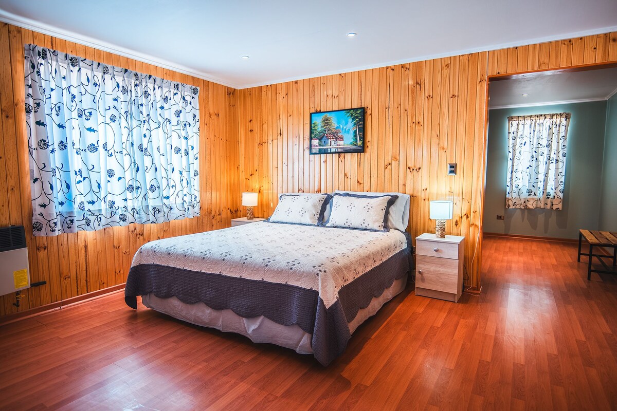 Un dormitorio con una pared revestida de madera, una cama con cubrecama floral, dos mesitas de noche con lámparas y un cuadro sobre la cama.