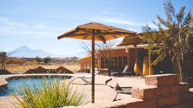 Zona de piscina con una sombrilla de madera, una tumbona y vistas a montañas al fondo.
