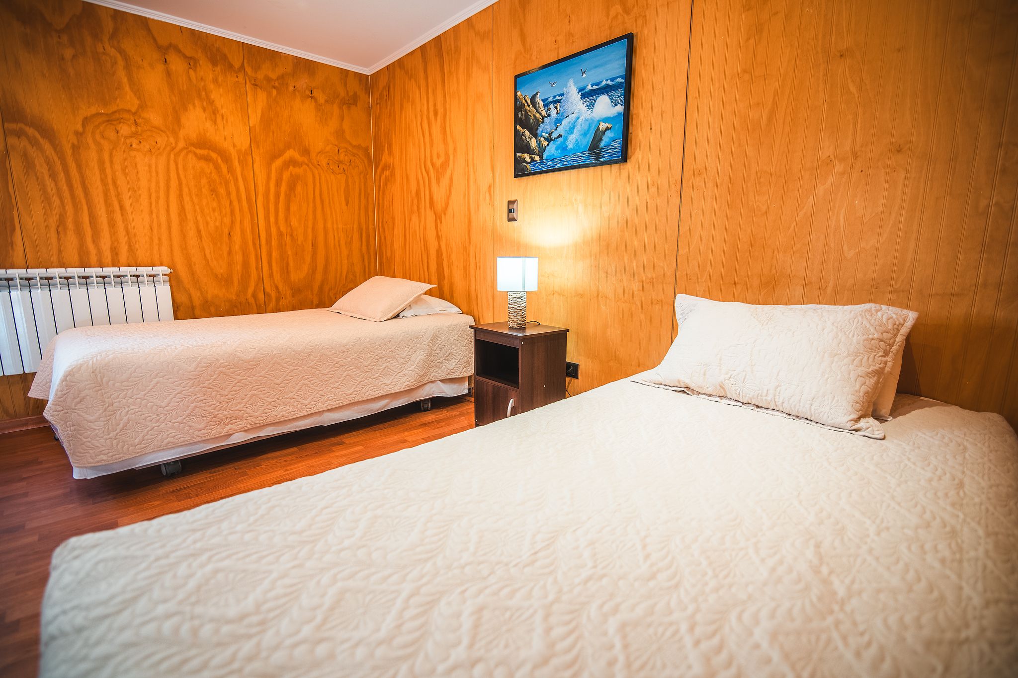 A hotel room with two beds, a wooden wall, a painting, and a nightstand with a lamp.