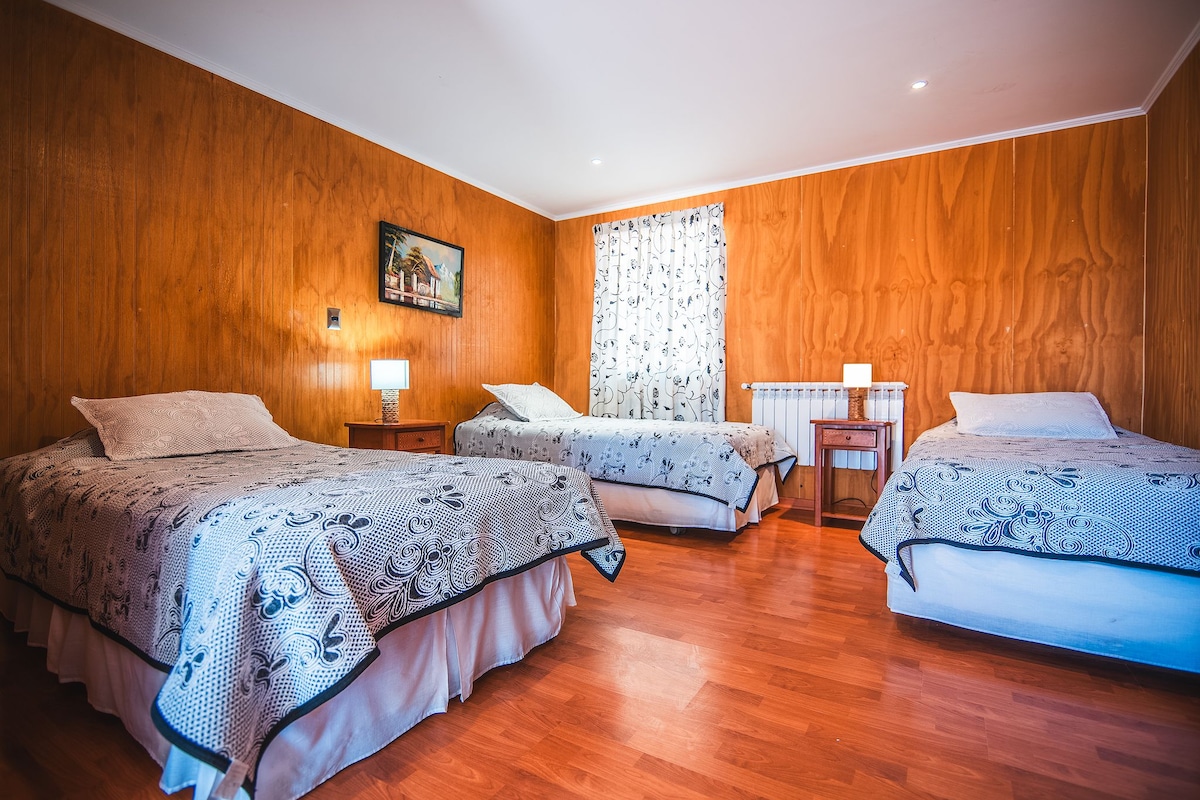 Habitación de hotel con tres camas, paredes de madera, un cuadro y una ventana con cortinas.