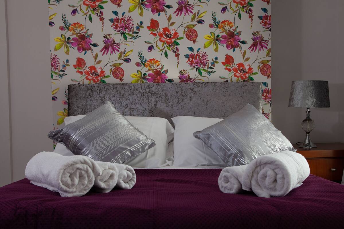 Una cama bien tendida con una colcha morada, dos almohadas y toallas enrolladas.