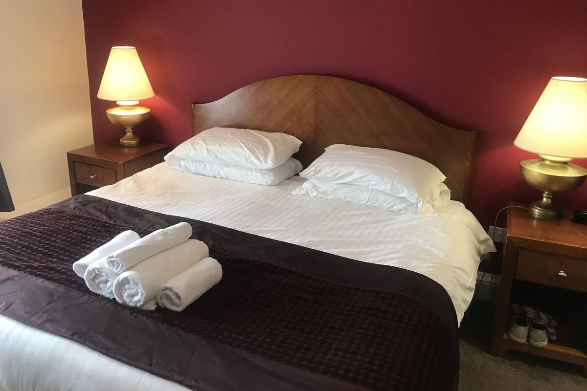 Una cama bien hecha con sábanas blancas y dos almohadas, un edredón marrón oscuro, y un juego de toallas sobre la cama. Hay dos mesitas de noche con lámparas a ambos lados de la cama.