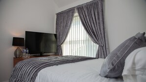 A neatly made bed with a striped blanket, a flat-screen TV on a wooden nightstand, and a window with sheer curtains.