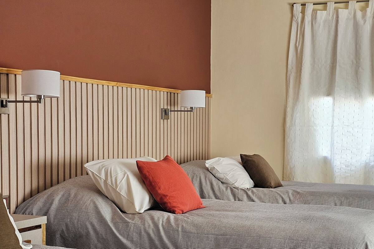 Una habitación de hotel con dos camas, ropa de cama a rayas, cabecera de madera, lámparas fijadas a la pared y una ventana con cortinas.