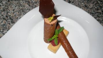 A dessert on a white plate with a chocolate cone, a square piece of cake, and a chocolate bar.