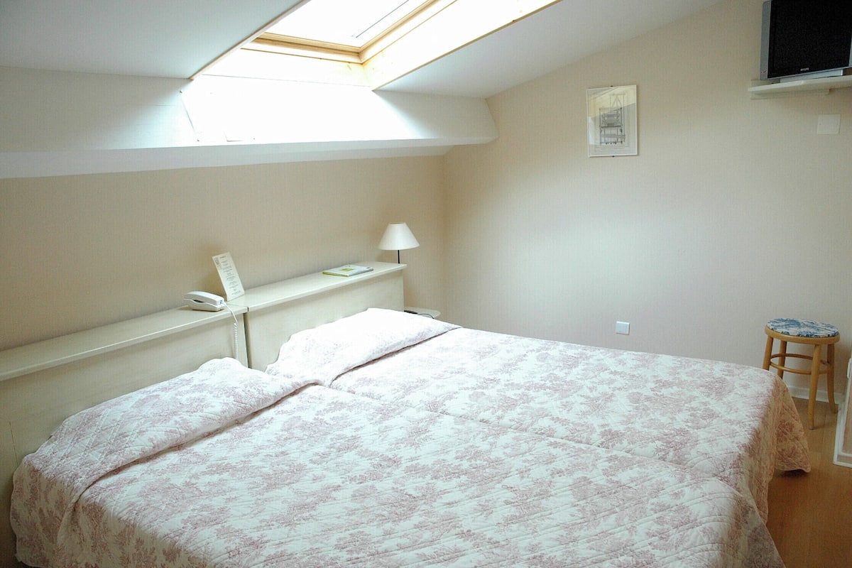 A bedroom with a bed, a bedside table with a lamp, a small stool, a window with a view, and a framed picture on the wall.