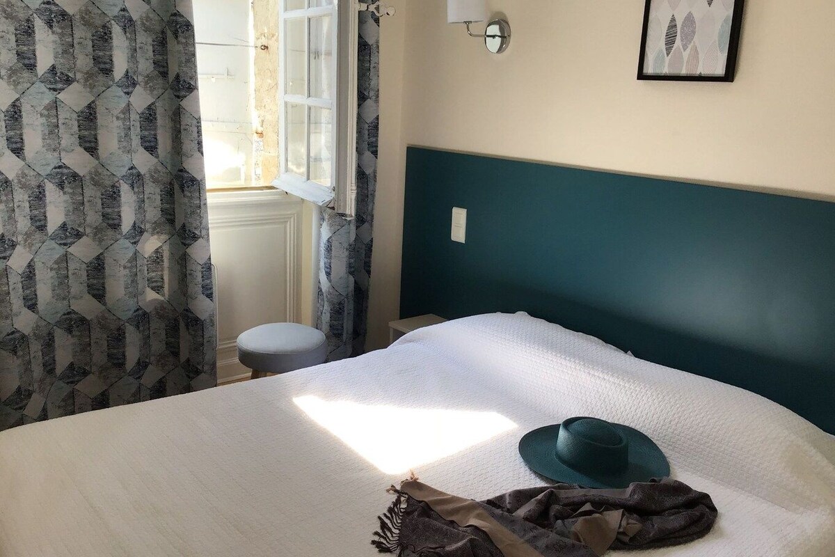 A bed with a white cover, a green headboard, a window with curtains, a wall-mounted light, and a framed picture.