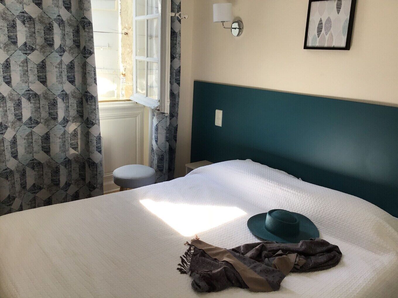 A bed with a white cover, a green headboard, a window with curtains, a wall-mounted light, and a framed picture.