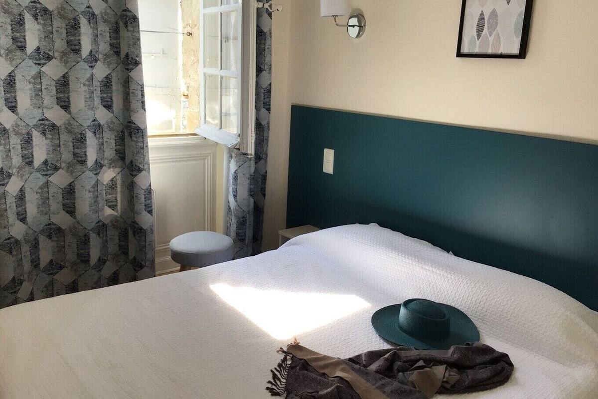 A bed with a white cover, a green headboard, a window with curtains, a wall-mounted light, and a framed picture.