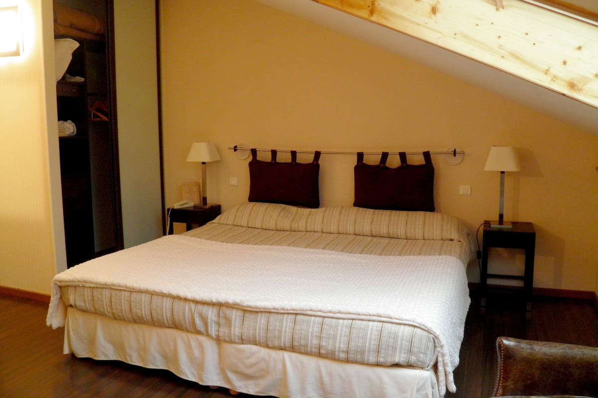 A bedroom with a large bed, two bedside tables with lamps, a wooden ceiling, and a wardrobe.