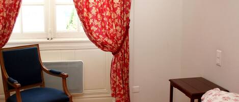 A bedroom with a bed, a chair, a table, and a window with red curtains.