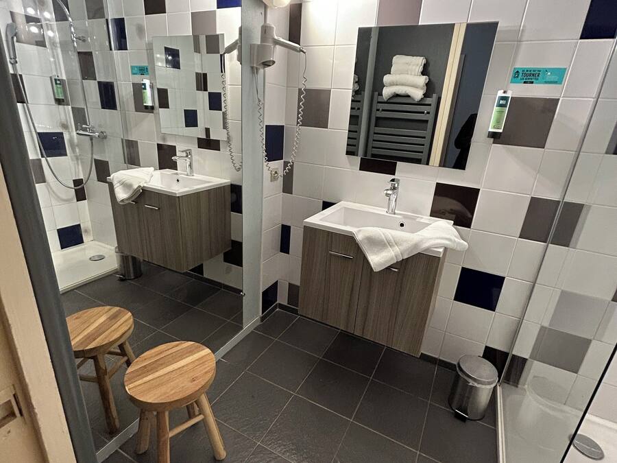 Un baño moderno con dos lavamanos, ducha y espejo. Hay taburetes de madera y un perchero para toallas.