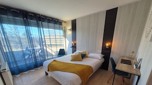 A modern hotel room with a large bed, a desk, and a chair. There is a view of the outdoors through the window. - Hotel Les Gravades (Ussel)
