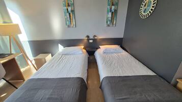 Habitación de hotel con dos camas individuales, un escritorio, una silla, una lámpara y dos obras de arte colgadas en la pared.