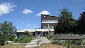 A modern building with a flat roof, a covered walkway, and a red spherical object on the ground. - Hotel Les Gravades (Ussel)