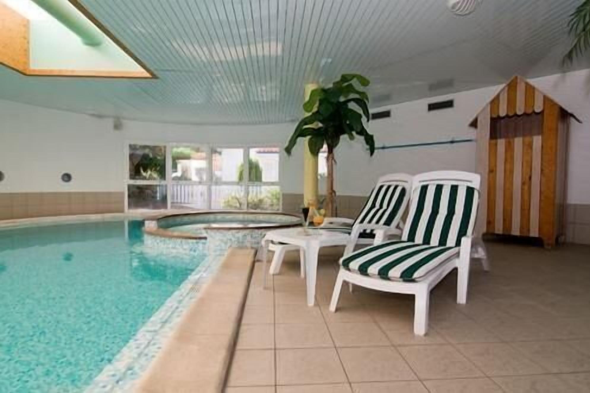 Une piscine intérieure avec un sol carrelé, deux chaises longues blanches à rayures vertes, une plante en pot et une petite structure en forme de cabane en bois.