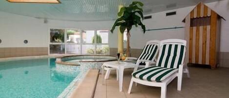 An indoor swimming pool with tiled flooring, two white lounge chairs with green stripes, a potted plant, and a small wooden hut structure.