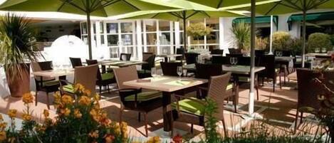 Un espacio de comedor al aire libre con mesas y sillas, sombrillas verdes y plantas en macetas.